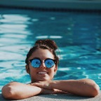 woman with sunglasses, in a pool, rests her arms on the ledge and smiles